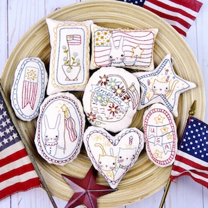 May include: A collection of patriotic-themed, hand-stitched fabric ornaments in various shapes, including stars, hearts, and pillows. Each ornament features red, white, and blue embroidery with American flag and animal designs. The ornaments are arranged on a wooden tray.