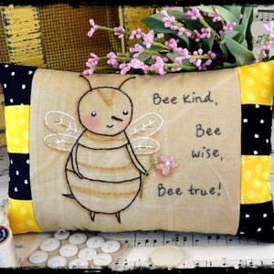 May include: A rectangular decorative pillow with a tan center panel featuring an embroidered bee and the text Bee Kind, Bee wise, Bee true! The pillow has black and yellow polka-dot side panels and is accented with a small pink flower applique.