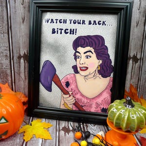 May include: A framed illustration featuring a person with dark purple hair wearing a pink floral top, holding a purple axe. The text WATCH YOUR BACK... BITCH! is printed in dark blue letters above the figure against a silver glitter background.