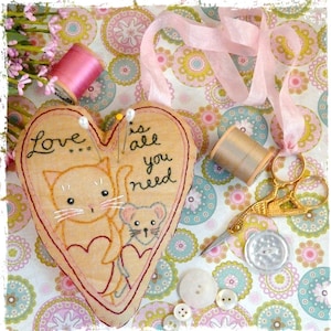Puede incluir: Un cojín de alfileres en forma de corazón con gatos bordados y el texto "Love is all you need". El cojín está sobre un fondo de tela con una cinta rosa, tijeras doradas y carretes de hilo.