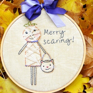 May include: A circular wooden embroidery hoop containing beige fabric with a hand-stitched design of a mummy figure holding a cat-shaped bucket. The text Merry scaring! is embroidered in black thread. A purple and lavender ribbon is tied at the top.