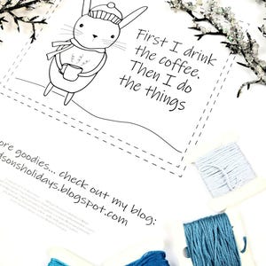 May include: A white print with a cartoon rabbit wearing a hat and scarf, holding a mug. Text reads "First I drink the coffee. Then I do the things." Several spools of blue embroidery floss are arranged around the print.