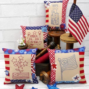 Peut inclure: Plusieurs coussins décoratifs patriotiques avec des broderies. Les coussins présentent des motifs rouges, blancs et bleus, des étoiles et des drapeaux américains. Certains coussins portent les inscriptions "BOOM!" et "AMERICA 1776-2026 250 YEARS". Des étoiles décoratives sont éparpillées.