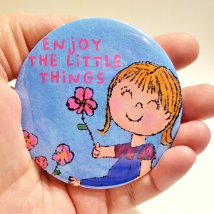 May include: A blue and pink round button with the text "Enjoy the little things" and a cartoon illustration of a girl with blonde hair holding a pink flower.