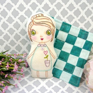 May include: A handmade doll with blonde hair, green eyes, and a light blue dress. The doll is holding a small flower in a pot. The doll is sitting on a checkered fabric.