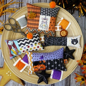 May include: Five Halloween-themed pillow ornaments with various patchwork designs. The pillows feature a black cat, a spiderweb, a mouse, a pumpkin, and a skeleton. The pillows are arranged on a wooden surface with spools of thread and other Halloween decorations.