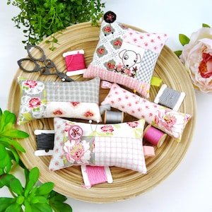 May include: A set of five fabric pincushions in various floral patterns, including a dog design. The pincushions are arranged on a woven basket with spools of thread and scissors.
