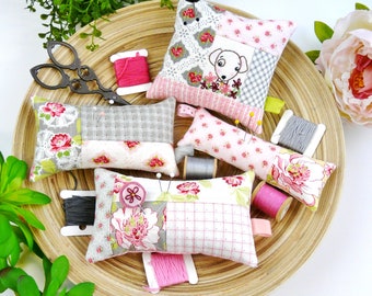 May include: A set of five fabric pincushions in various floral patterns, including a dog design. The pincushions are arranged on a woven basket with spools of thread and scissors.
