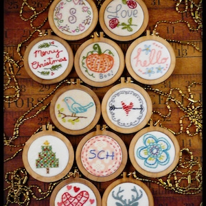 May include: Twelve small, round, wooden embroidery hoops with various colorful embroidery designs. The designs include a Christmas tree, a pumpkin, a heart with an arrow, a bird, a flower, a deer, a heart, a tree with a star, a wreath, the words "Merry Christmas", "Love", "Hello", "Boo", and "SCH".