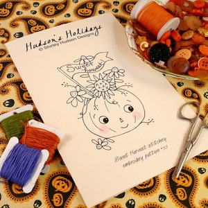 May include: A black and white line drawing of a pumpkin with a smiling face and a banner that says "Sweet Harvest!"  The drawing is on a white card with the text "Hudson's Holidays © Shirley Hudson Designs" and "Sweet Harvest stitchery embroidery pattern - 501".