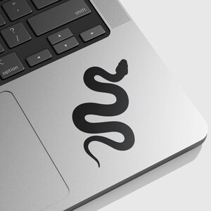 May include: A matte black vinyl sticker featuring a simple, solid silhouette of a snake in a winding, S-shaped posture. The sticker is applied to the silver metallic surface of a laptop, positioned near the keyboard and trackpad area.