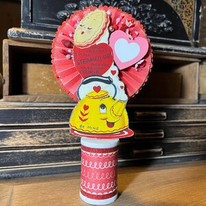 May include: A vintage Valentine's Day decoration featuring a red and white paper rosette with a heart and a yellow teapot with the text "Gosh I'm all Steamed Up over you you're some cookie" and "Be Mine".