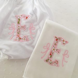 May include: White baby bloomers and a matching burp cloth, both embroidered with a floral "E" in pink and green, accented with pink bows. The bloomers have a ruffled edge.