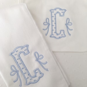 May include: Two white burp cloths with blue embroidered letters and bows. The letter "C" is embroidered on each cloth.