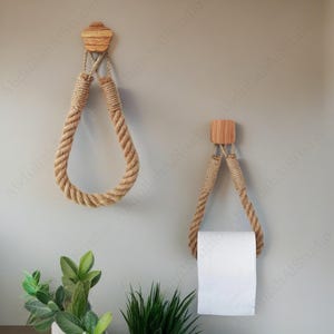 May include: Two rustic rope and wood bathroom accessories. One is a towel holder, and the other is a toilet paper holder. The rope is a natural beige colour, and the wood is light brown. The holders are mounted on a light grey wall.