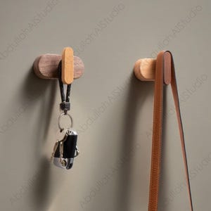 Puede incluir: Dos ganchos de pared de madera con una correa de cuero marrón colgando de uno. El gancho de la izquierda tiene un juego de llaves colgando de él.