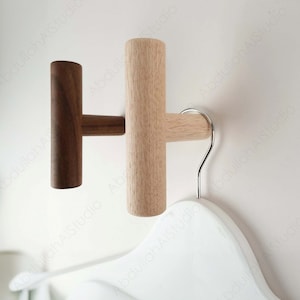 May include: Two wooden wall hooks, one light brown and one dark brown, with a T-shaped design. The hooks are mounted on a white wall and a white hanger is hanging on one of the hooks.