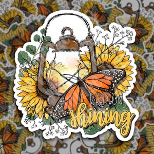 Pode incluir: Um autocolante com uma lanterna vintage, girassóis e uma borboleta monarca. A borboleta tem asas laranja e pretas. O texto "Keep on Shining" está em escrita amarela. Folhas verdes e flores brancas rodeiam a lanterna.