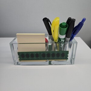 Könnte beinhalten: Ein durchsichtiger Acryl-Schreibtisch-Organizer mit einem Computer-RAM-Riegel und einem Stapel Haftnotizen. Der Organizer enthält Stifte und Marker in Schwarz, Gelb, Grün und Blau. Das Design ist modern und funktional.