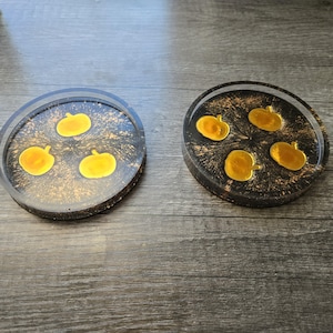 May include: Two round coasters with a black and gold speckled design. Each coaster features several yellow pumpkin shapes. The coasters are made of resin and are suitable for autumn or Halloween decor.
