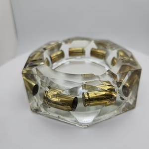 May include: A hexagonal ashtray made of clear resin with multiple brass-colored metallic bullet casings embedded throughout the structure. The transparent material allows for a clear view of the gold-toned casings arranged in a circular pattern.