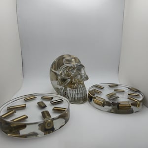 May include: A clear resin skull and two oval ashtrays with embedded brass bullet casings. The skull is positioned between the ashtrays. The ashtrays are filled with various sizes of brass bullet casings. The resin has a slight amber tint.