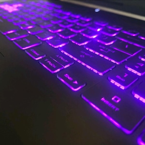 Op de afbeelding: Een laptoptoetsenbord met zwarte toetsen en heldere paarse achtergrondverlichting. De toetsen zijn duidelijk gemarkeerd met witte tekst, waaronder functietoetsen zoals Shift, Enter, PgUp, PgDn, End en Insert, die allemaal oplichten in paars.