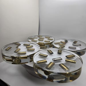 May include: A set of four circular coasters made of clear resin, each embedded with multiple metallic bullet casings in brass and silver tones. The coasters have a smooth, transparent finish that displays the cylindrical casings arranged inside.