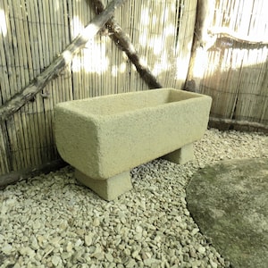 May include: A rectangular, light beige stone planter with rounded corners and a textured surface. The planter sits on two short, rectangular pedestals. The background features a bamboo fence and small gravel.