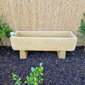 May include: A light brown stone trough planter with two short legs. The planter is sitting on a bed of brown mulch.