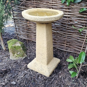May include: A yellow bird bath with a square base and a round bowl. The bird bath is made of a textured material and is sitting on a bed of brown mulch.
