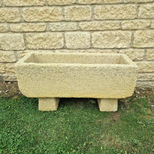 May include: A rectangular, light-yellow stone planter with a textured surface. The planter is supported by four short, rectangular feet. The background features a light-yellow brick wall and green grass.