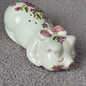 May include: White ceramic piggy bank with pink bow and purple flower design. The piggy bank is lying on its side with its eyes closed.