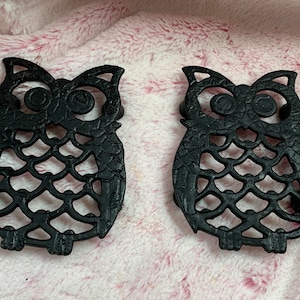 May include: Two black cast iron owl-shaped hot pads. The owls have a detailed design with cutouts and a textured surface.
