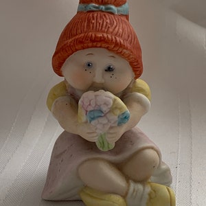 May include: A porcelain figurine of a girl with red hair, wearing a yellow and pink dress, holding a bouquet of flowers. She has one blue eye and one closed eye, and is sitting with her legs crossed.