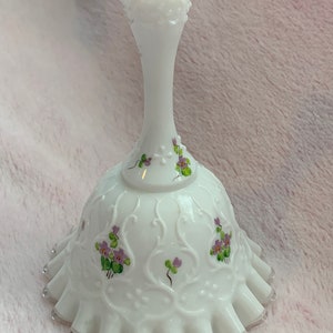 May include: A white milk glass bell with a scalloped edge and a delicate floral design featuring purple and green violets. The bell has a white handle with a scalloped top.