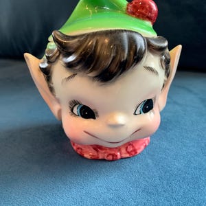 Peut inclure: Une figurine en céramique représentant la tête d'un lutin avec un chapeau vert et un pompon rouge. Le lutin a les cheveux bruns, les yeux bleus et une écharpe rose.