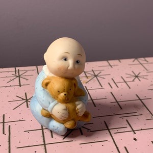May include: A small ceramic figurine of a bald baby in blue pyjamas holding a brown teddy bear.