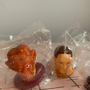 Puede incluir: Tres peque&ntilde;as figuras de pl&aacute;stico en bolsas de pl&aacute;stico transparentes individuales. Las figuras son de un alien&iacute;gena marr&oacute;n con orejas grandes, un hombre con cabello casta&ntilde;o y un alien&iacute;gena marr&oacute;n con orejas grandes. Las figuras est&aacute;n sobre una superficie a cuadros rosa y blanca.