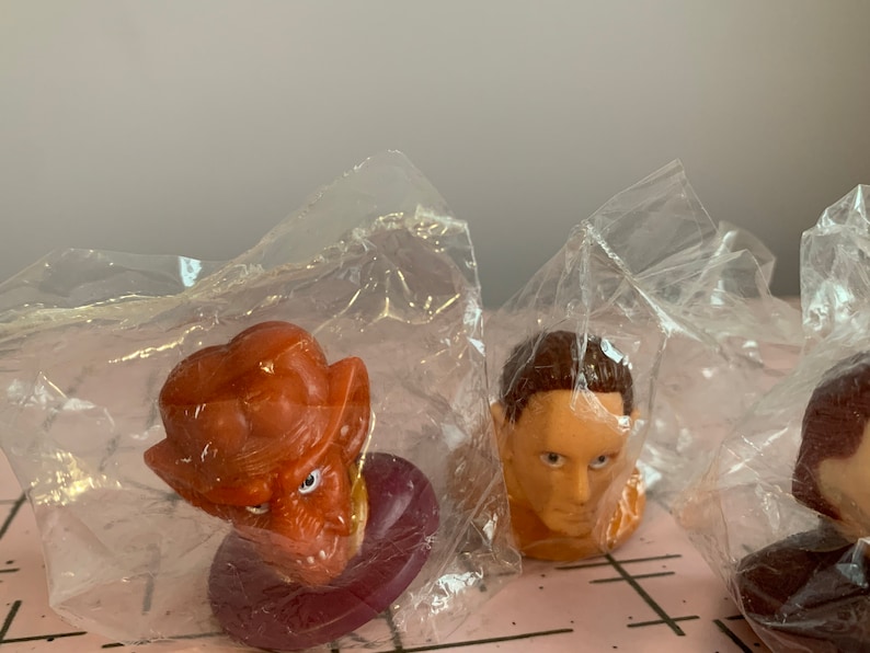 Puede incluir: Tres peque&ntilde;as figuras de pl&aacute;stico en bolsas de pl&aacute;stico transparentes individuales. Las figuras son de un alien&iacute;gena marr&oacute;n con orejas grandes, un hombre con cabello casta&ntilde;o y un alien&iacute;gena marr&oacute;n con orejas grandes. Las figuras est&aacute;n sobre una superficie a cuadros rosa y blanca.