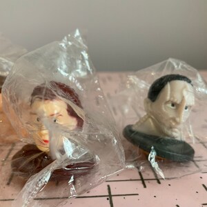 Puede incluir: Tres figuras de pl&aacute;stico de cabezas de personas famosas, empaquetadas en bolsas de pl&aacute;stico transparentes. Las figuras son de un hombre con cabello casta&ntilde;o, una mujer con cabello casta&ntilde;o y un hombre con cabello negro.