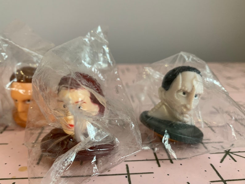 Puede incluir: Tres figuras de pl&aacute;stico de cabezas de personas famosas, empaquetadas en bolsas de pl&aacute;stico transparentes. Las figuras son de un hombre con cabello casta&ntilde;o, una mujer con cabello casta&ntilde;o y un hombre con cabello negro.