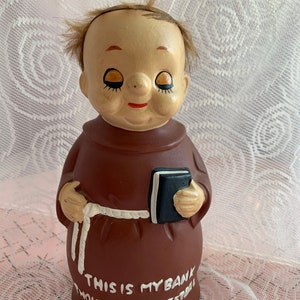 Puede incluir: Una alcancía de cerámica marrón con forma de monje que sostiene un libro. La alcancía tiene un cinturón de cuerda blanco y las palabras "This is my bank Thou shalt not steal" escritas en la parte inferior.