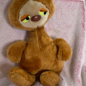 Può includere: Un giocattolo di peluche marrone con un collo lungo e grandi occhi tristi. Il giocattolo è seduto su una coperta rosa pelosa.