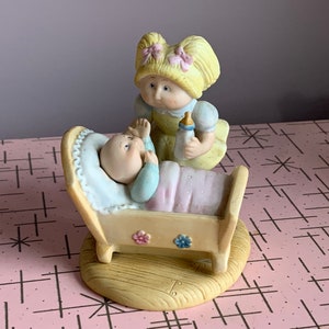 Peut inclure: Une figurine en porcelaine d'une fille aux tresses blondes tenant un biberon, debout à côté d'un berceau avec un bébé à l'intérieur. La figurine est peinte dans des couleurs pastel et a un style fantaisiste.