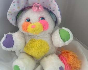 May include: White plush toy with a pink, yellow, green, and purple color scheme. The toy is wearing a purple and white star-patterned hat. The toy has a pink and orange fluffy tail.