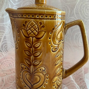 Puede incluir: Una jarra de cerámica marrón con tapa y asa. La jarra tiene un diseño floral con corazones y hojas.