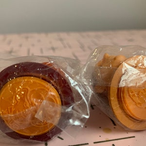 Puede incluir: Dos peque&ntilde;os juguetes redondos de pl&aacute;stico naranja y marr&oacute;n envueltos en pl&aacute;stico transparente. Los juguetes tienen un dise&ntilde;o en relieve en la parte superior y un anillo rojo alrededor de la parte inferior.