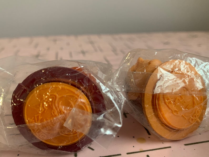 Puede incluir: Dos peque&ntilde;os juguetes redondos de pl&aacute;stico naranja y marr&oacute;n envueltos en pl&aacute;stico transparente. Los juguetes tienen un dise&ntilde;o en relieve en la parte superior y un anillo rojo alrededor de la parte inferior.