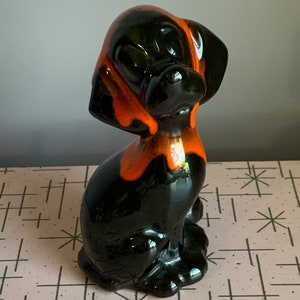 Puede incluir: Una figurita de perro de cerámica negra y naranja. El perro está sentado con la cabeza inclinada hacia un lado. La figurita está esmaltada en un esquema de colores negro y naranja.